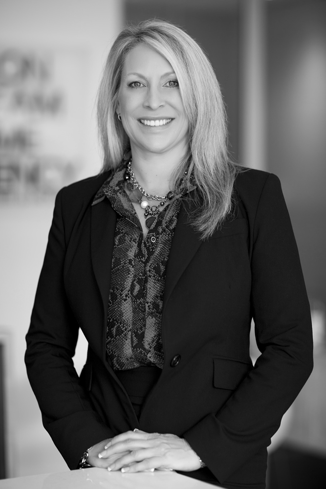 Black and white Headshot of a female wearing a dark suit and standing in an office environment in Auckland New Zealand. Captured by professional Auckland photographer Adam Firth