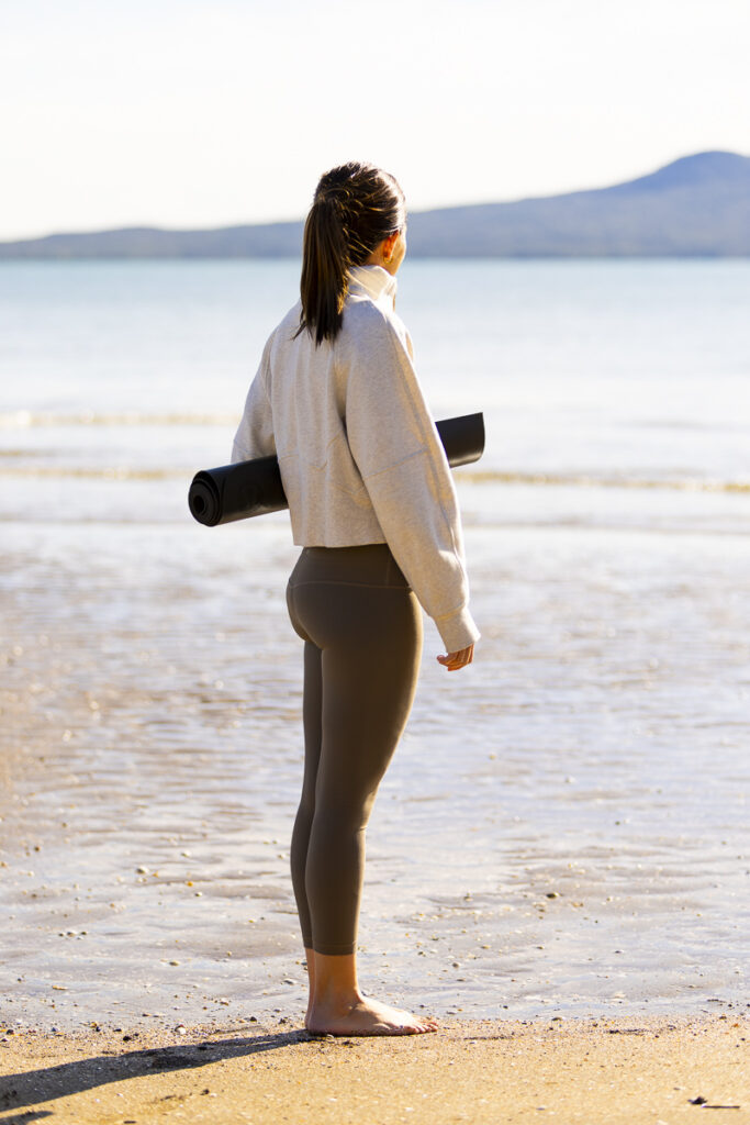 Woman looking out to Rangitoto island from Saint Heliers beach holding a yoga mat. Photographed by Adam Firth