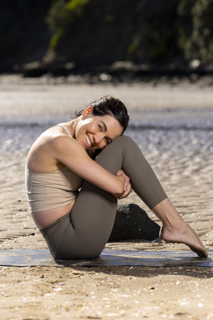 Professional photograph of a woman on a yoga mat smiling at camera while hugging her knees photographed by local photographer Adam Firth at Saint Heliers beach in Auckland.