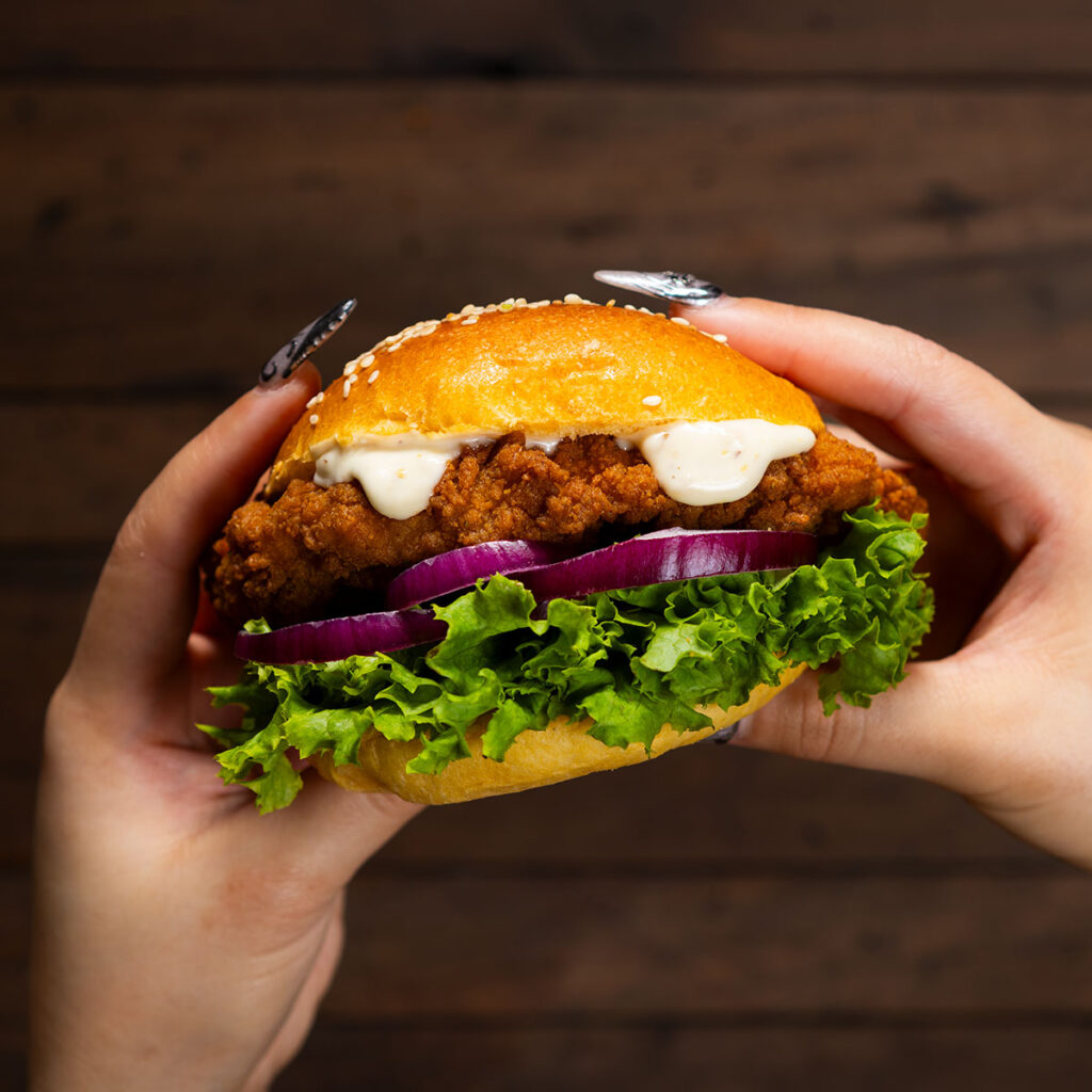 hands holding a chicken burger photographed over a dark wooden background by professional photographer Adam Firth